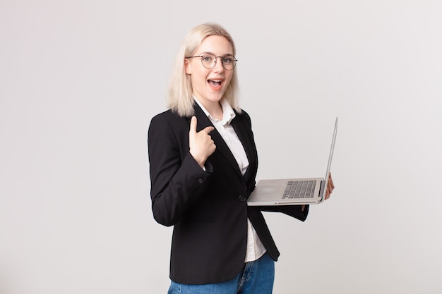 Blond pretty woman feeling happy and pointing to self with an excited and holding a laptop