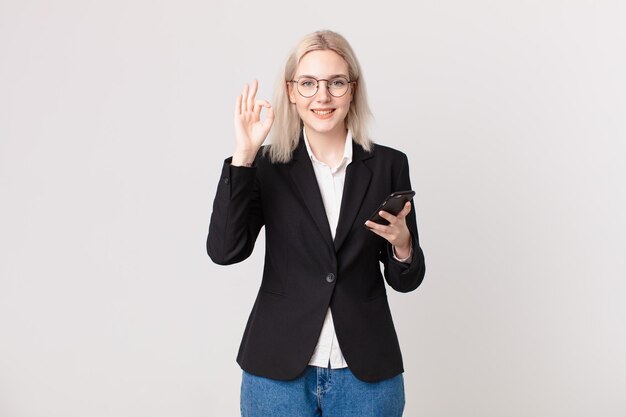 Blond pretty woman feeling happy, showing approval with okay gesture and holding a mobile telephone