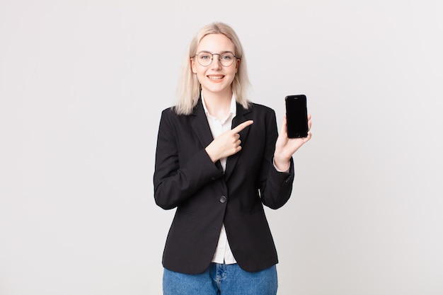 Bella donna bionda che sorride allegramente, si sente felice e indica un lato e tiene in mano un telefono cellulare