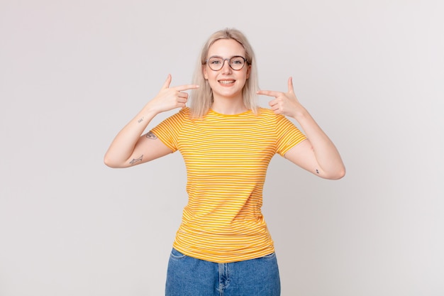 Blond pretty woman smiling confidently pointing to own broad smile