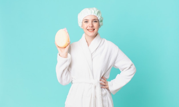 Blond pretty woman smiling happily with a hand on hip and confident and wearing bathrobe