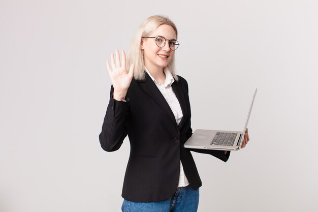 Blond pretty woman smiling and looking friendly, showing number five and holding a laptop