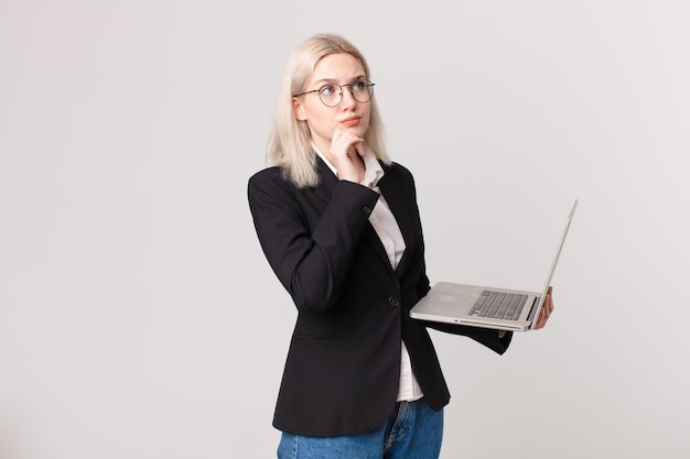 Blond pretty woman thinking, feeling doubtful and confused and holding a laptop