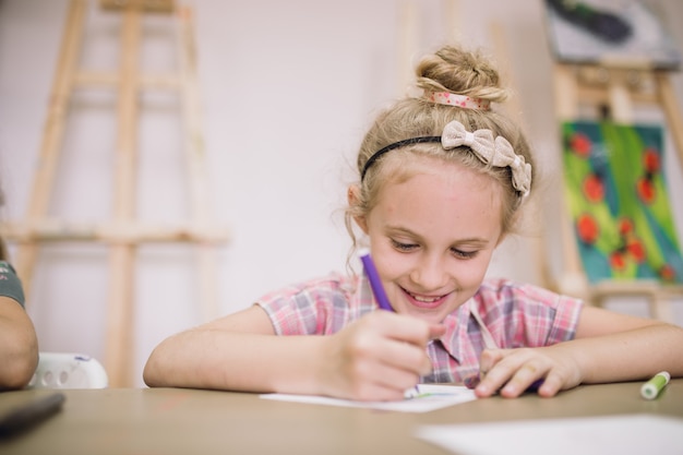 Blond schattig glimlachend zevenjarig meisje, tekent aan de tafel in de creatieve studio.