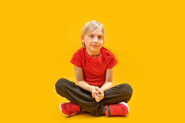Photo blond schoolboy wears red tshirt and sneakers sit on the floor with crossed legs teenager yellow background
