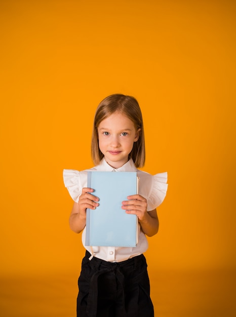Blond schoolmeisje in uniform houdt een blauw notitieboekje op een gele achtergrond vast met een kopie van de spatie