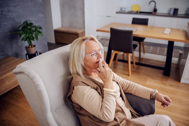 Blond senior woman sitting in chair at home and spraying nose spray.