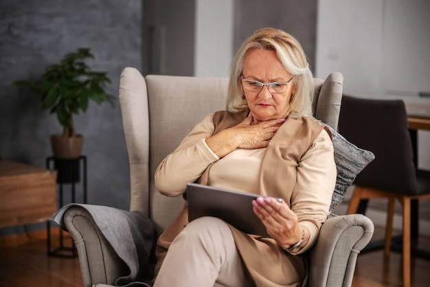 Senior donna bionda seduta a casa, avendo dolore nel pensiero e guardando tablet per ottenere consigli online.