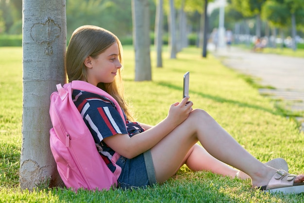 Ragazza bionda del bambino dello studente con lo smartphone in parco