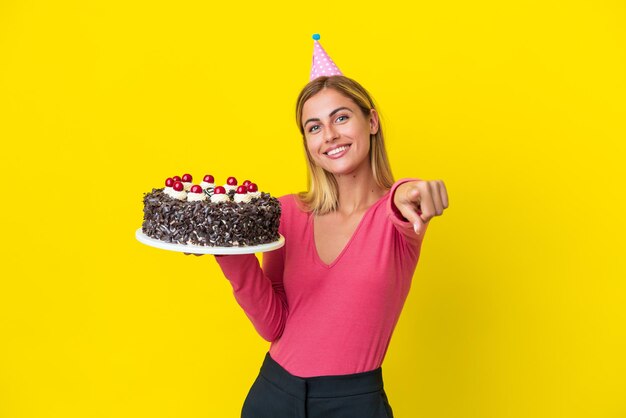 Blond Uruguayaans meisje met verjaardagstaart geïsoleerd op gele achtergrond wijzend naar voren met gelukkige uitdrukking