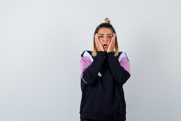 Blond woman in black tracksuit holding cheeks with palms and looking surprised