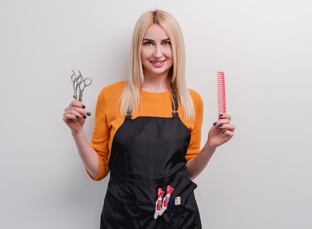 Photo blond woman hairdresser smiling and holding in her hands professional scissors and comb isolated on white surface