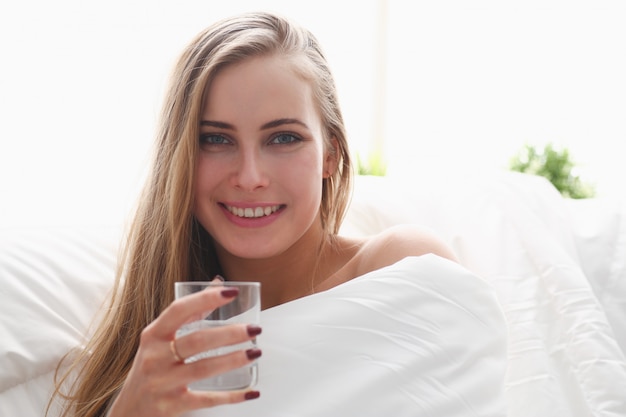 blond woman hold glass of water in arm early morning stay in bed