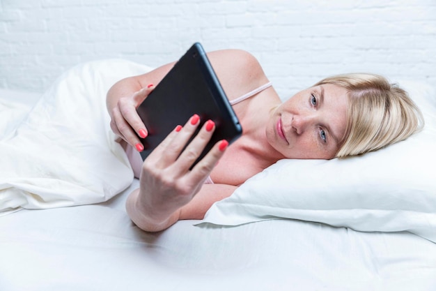 A blond woman is lying in bed with a tablet Messenger social networks communication Background from a white brick wall