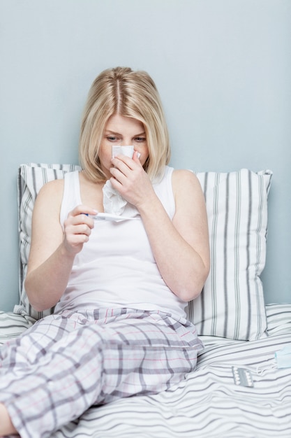 A blond woman is sitting in bed and looking at a thermometer wiping her nose with a handkerchief. Infectious diseases. Quarantine during the coronavirus pandemic. Vertical.