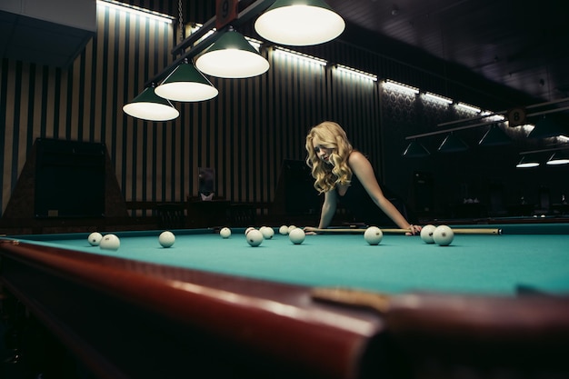 Блондинка играет, наслаждаясь бильярдными шарами на столе с зеленой поверхностью в бильярдном клубе