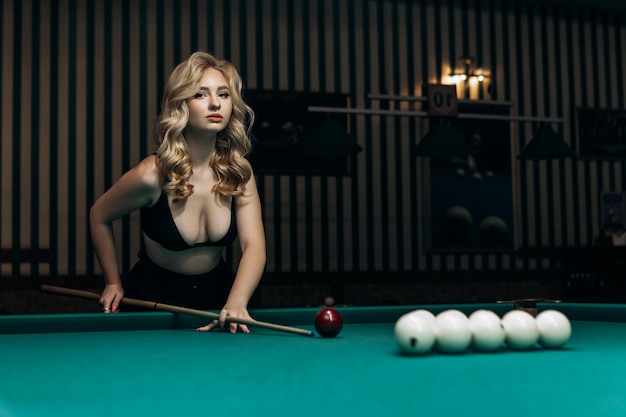 Blond woman playing enjoying billiard hold cue hit white billiard balls on table with green surface