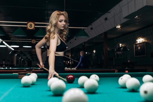 Blond woman playing enjoying billiard hold cue hit white billiard balls on table with green surface