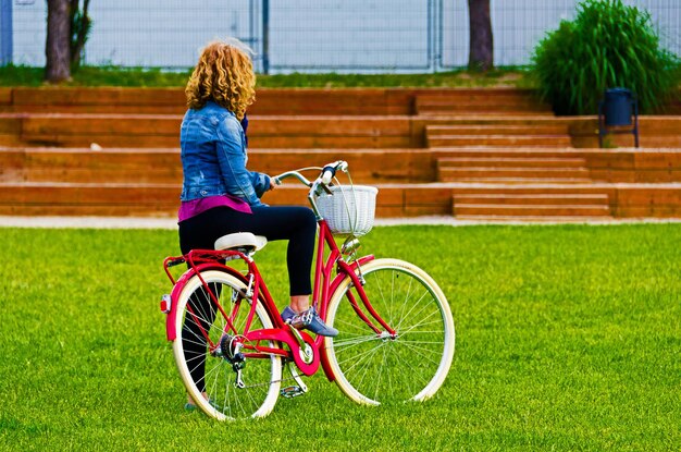 赤い自転車に青いデニムで公園を歩いている金髪の女性