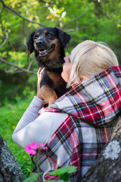 写真 緑の自然の中で腕を組んで彼女の犬を保持しているフード付きの金髪の女性。