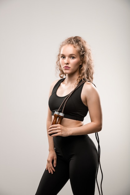 Blond young woman in activewear holding skipping-rope on her shoulder having break after training