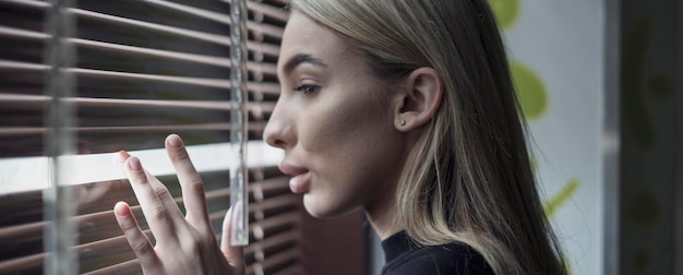 A blond young woman standing near the window and looking out through jalousie