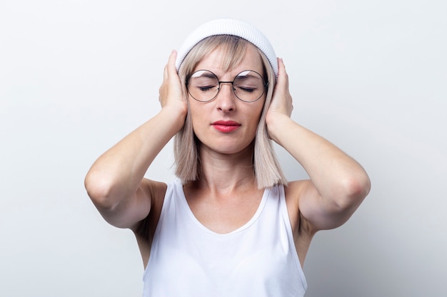 Blond young woman with closed eyes, covering her ears with her palms on a light background.