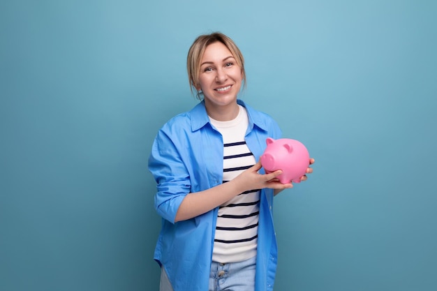 Blonde aantrekkelijke jonge vrouw in casual kleding die bedachtzaam een spaarvarken met geld op een blauw houdt