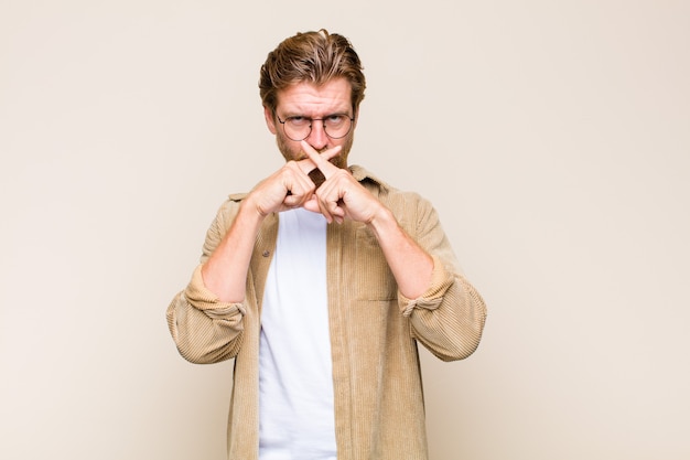 Blonde adult caucasian man looking serious and displeased with both fingers crossed up front in rejection, asking for silence