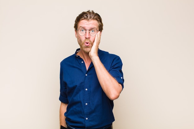 Blonde adult man feeling shocked and astonished holding face to hand in disbelief with mouth wide open