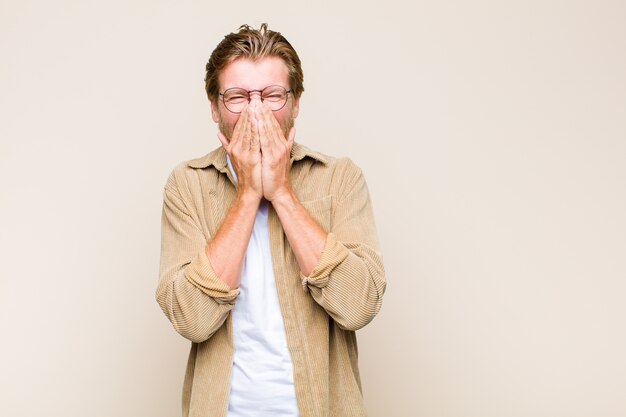 Blonde adult man looking happy, cheerful, lucky and surprised covering mouth with both hands