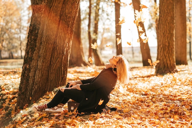 Blonde in autumn clothes enjoys life and puts her face under fall of leaves sitting on fallen leaves in city park. Fun while hiking in autumn season. Lifestyle. Colors of autumn.