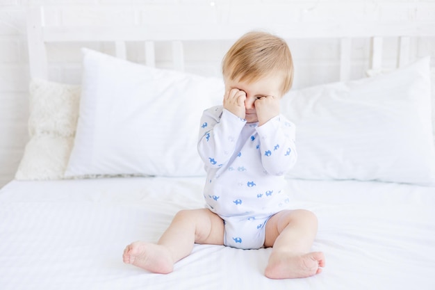 A blonde baby boy rubs sore inflamed eyes or wants to sleep on a white crib while sitting in a room at home with white cotton bedding the concept of childrens goods and accessories