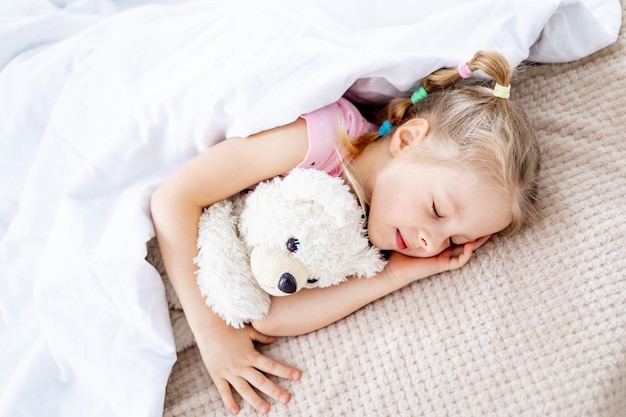 a blonde baby girl is sleeping sweetly under a white blanket holding a teddy bear in her hands on a white bed in a bright bedroom