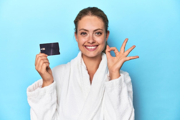 Blonde in bathrobe with credit card in blue studio cheerful and confident showing ok gesture