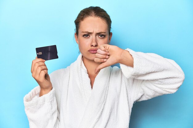 Blonde in bathrobe with credit card in blue studio showing a dislike gesture thumbs down