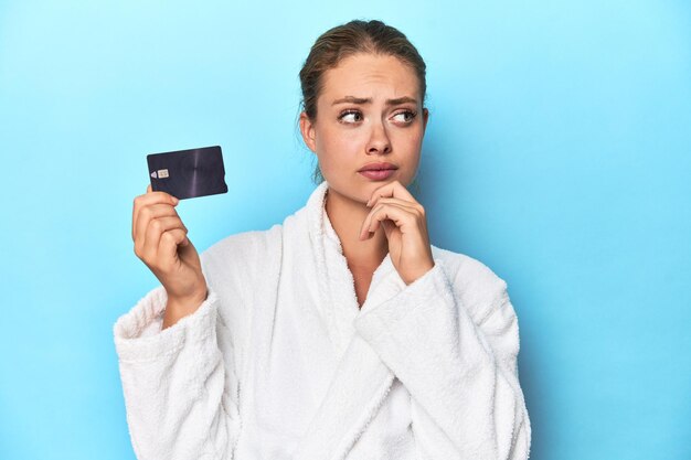 Photo blonde in bathrobe with credit card looking sideways with doubtful and skeptical expression