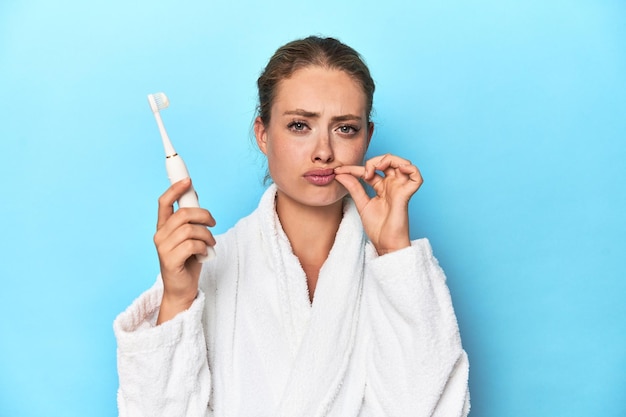 Blonde in bathrobe with electric toothbrush in studio with fingers on lips keeping a secret