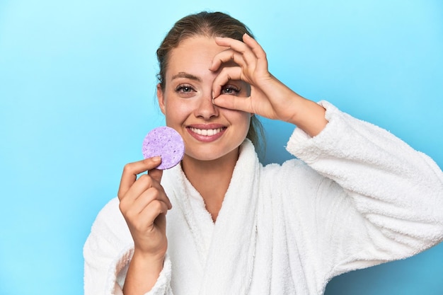 Blonde in bathrobe with facial sponge in studio excited keeping ok gesture on eye