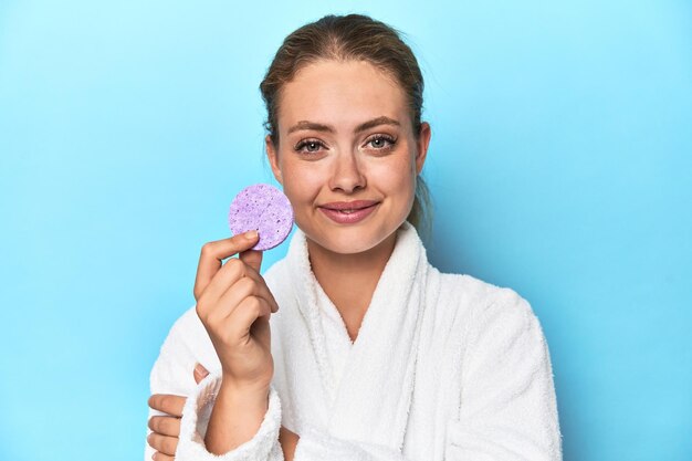 Blonde in bathrobe with facial sponge in studio happy smiling and cheerful