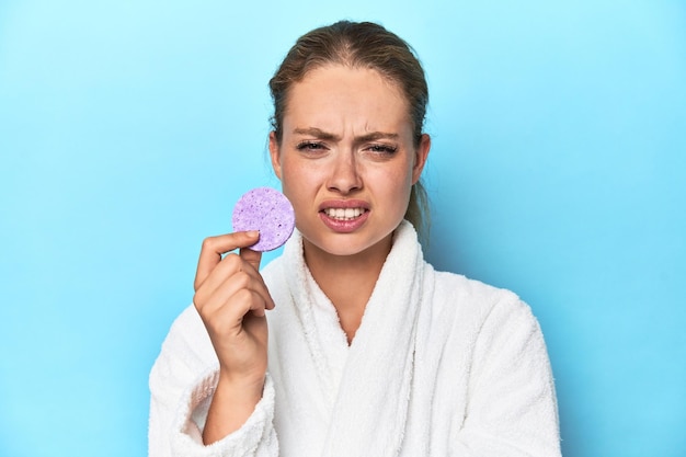 Blonde in bathrobe with facial sponge in studio screaming very angry and aggressive