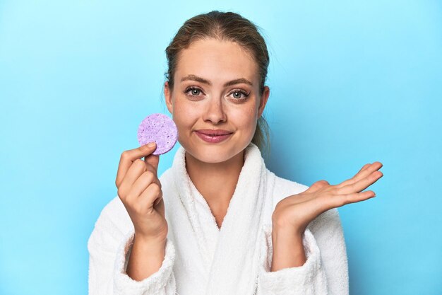 Blonde in bathrobe with facial sponge in studio showing a copy space on a palm