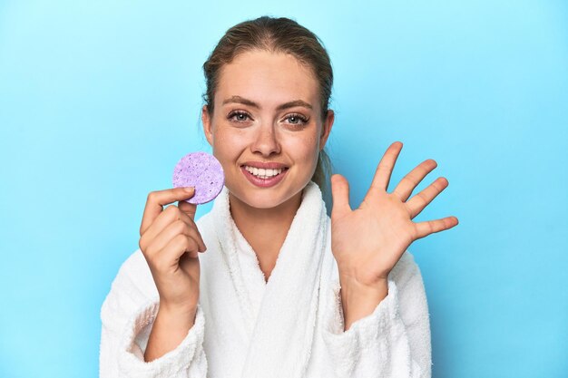 Blonde in bathrobe with facial sponge in studio smiling cheerful showing number five with fingers