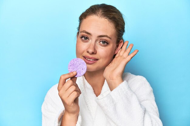 Blonde in bathrobe with facial sponge in studio trying to listening a gossip