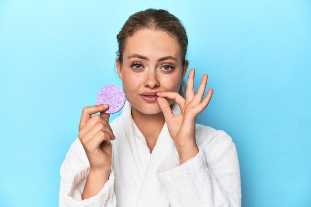 Blonde in bathrobe with facial sponge in studio with fingers on lips keeping a secret