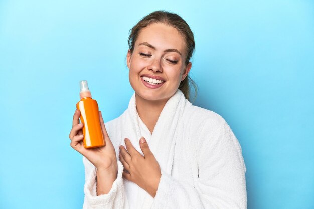 Blonde in bathrobe with sunscreen cream in studio laughs out loudly keeping hand on chest