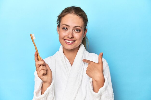 Blonde in bathrobe with a toothbrush in blue studio person pointing by hand to a shirt copy space