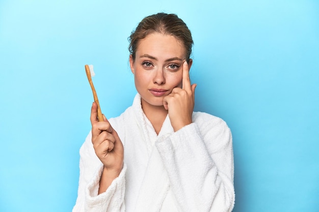 Blonde in bathrobe with a toothbrush in blue studio pointing temple with finger thinking focused