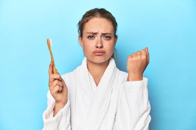 Blonde in bathrobe with a toothbrush in blue studio showing fist to camera aggressive