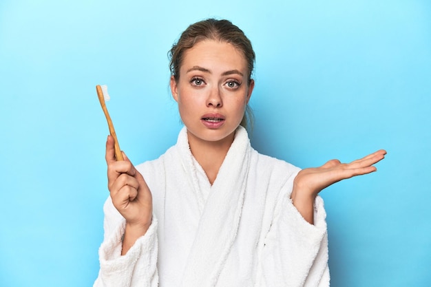 Blonde in bathrobe with a toothbrush in blue studio surprised and shocked
